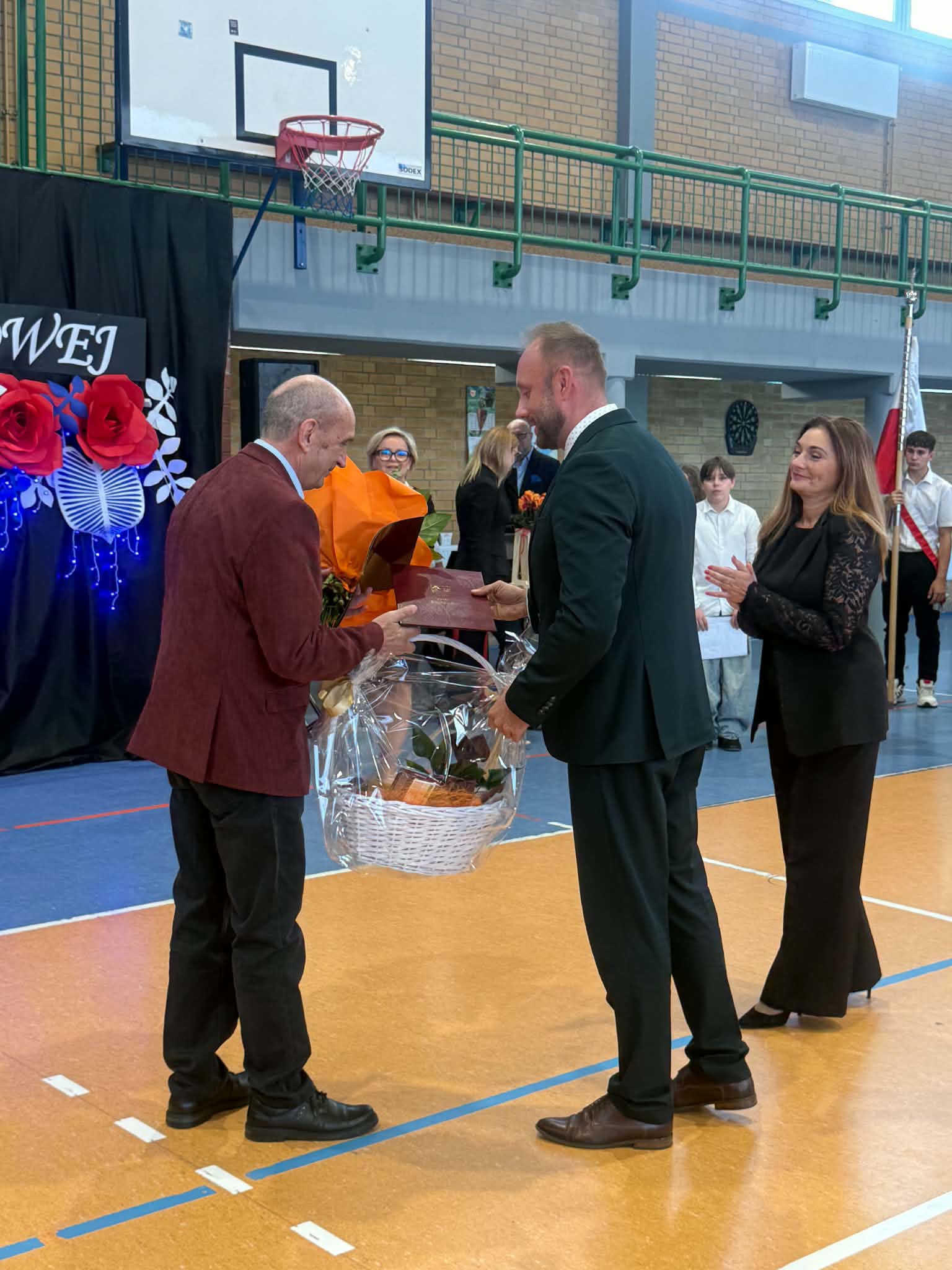 Mężczyzna wręcza kosz upominkowy oraz&nbsp;dyplom drugiemu mężczyźnie podczas uroczystości w&nbsp;hali sportowej