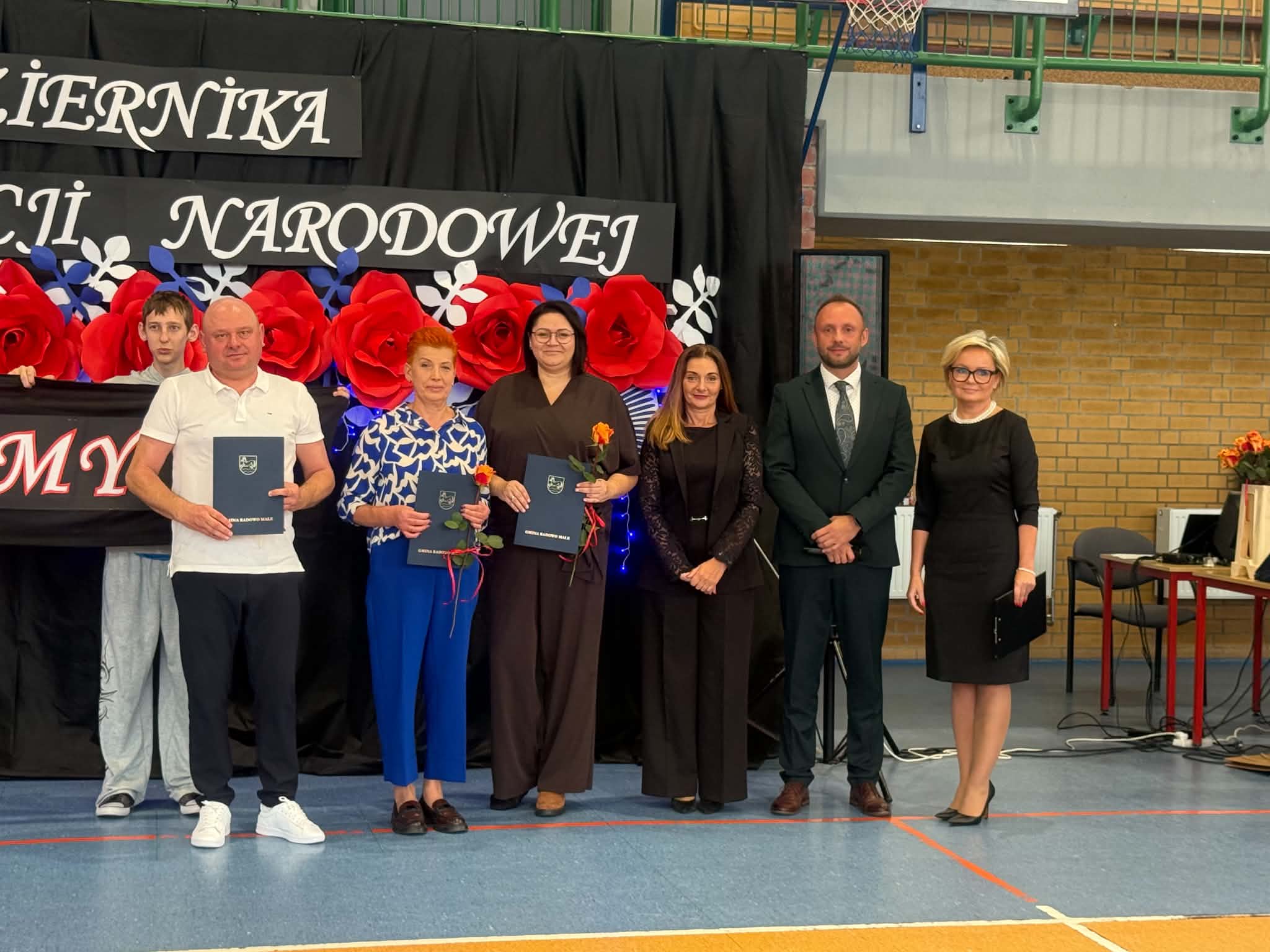 Grupa os&oacute;b stojąca na&nbsp;tle dekoracji z&nbsp;okazji Dnia Edukacji Narodowej, trzymająca dyplomy i&nbsp;kwiaty.