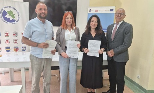 Cztery osoby prezentują podpisane dokumenty podczas oficjalnego spotkania. W tle widoczne są banery programu Fundusze Europejskie oraz Związku Zachodniopomorskiej Strefy Centralnej.