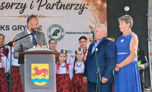 Na scenie podczas lokalnej uroczystości stoi kilka osób. Przy mównicy z herbem Gminy Radomyś Mate przemawia mężczyzna w szarym garniturze. Obok niego stoją starszy mężczyzna w granatowym garniturze i kobieta w niebieskiej sukience. W tle widać dzieci i kobiety w tradycyjnych strojach ludowych