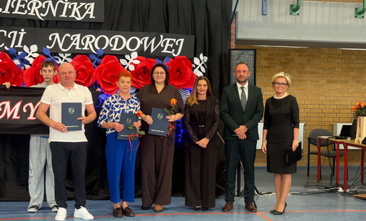 Grupa osób stojąca na tle dekoracji z okazji Dnia Edukacji Narodowej, trzymająca dyplomy i kwiaty.